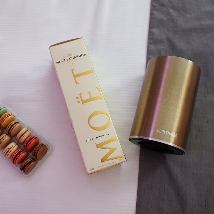 The coolenator gold design wine cooler on a bed on the right side, in the middle a box of champagne moet and on the right a box with macarons.