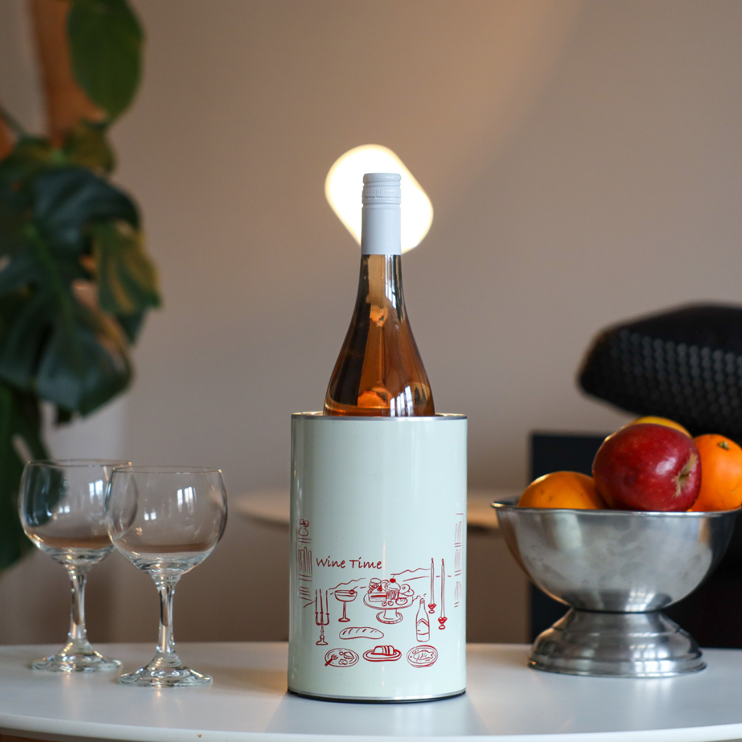 The Coolenator wine cooler with wine time design in the middle of the white table with a bottle of rose wine in it. On the left side two glasses of wine and on the right side a bowl with fruit. Situated in a home. 