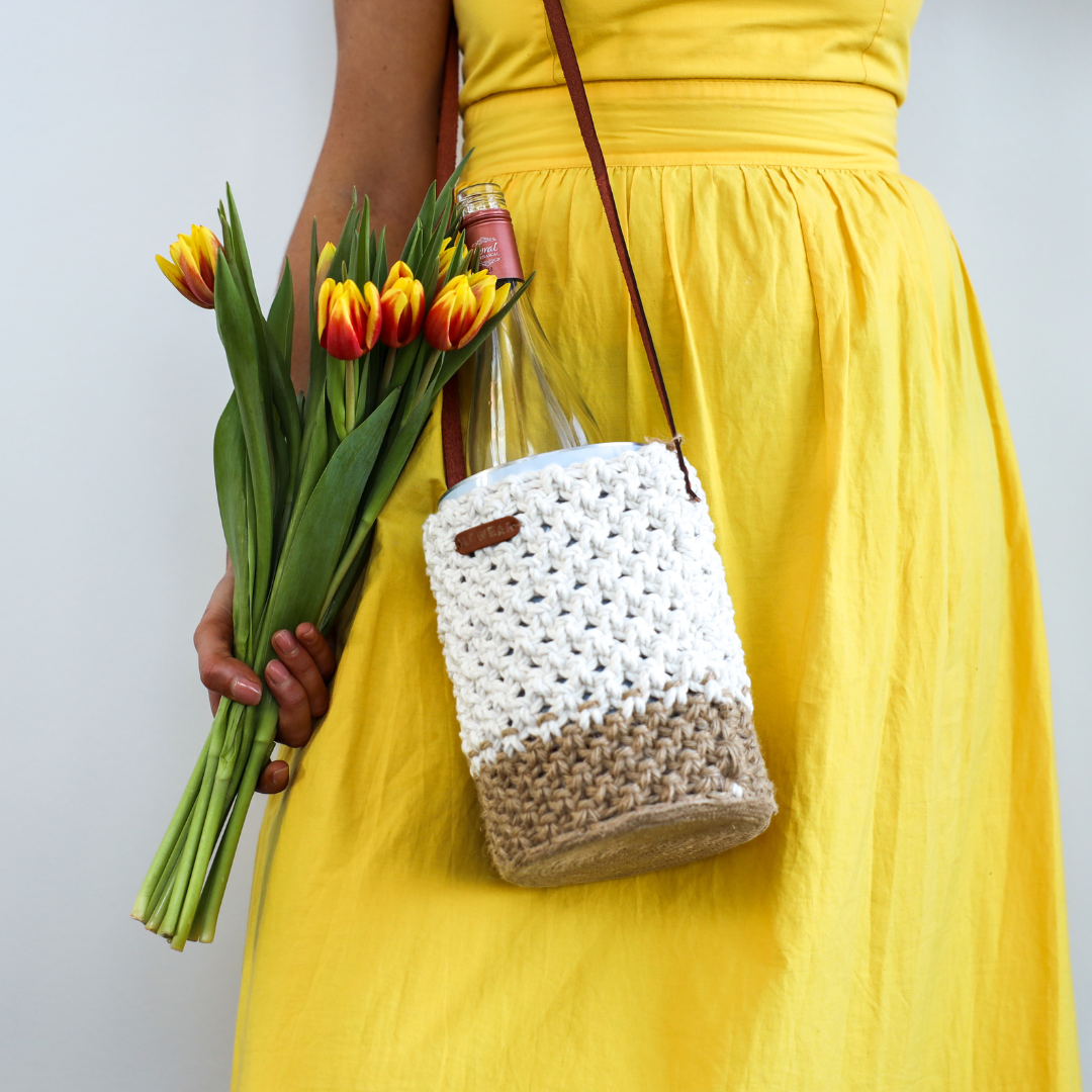 A woman in a yellow dress holding tulips and wearing a withe and beige crochet bag with leather straps with a coolenator wine cooler and bottle of wine in it.