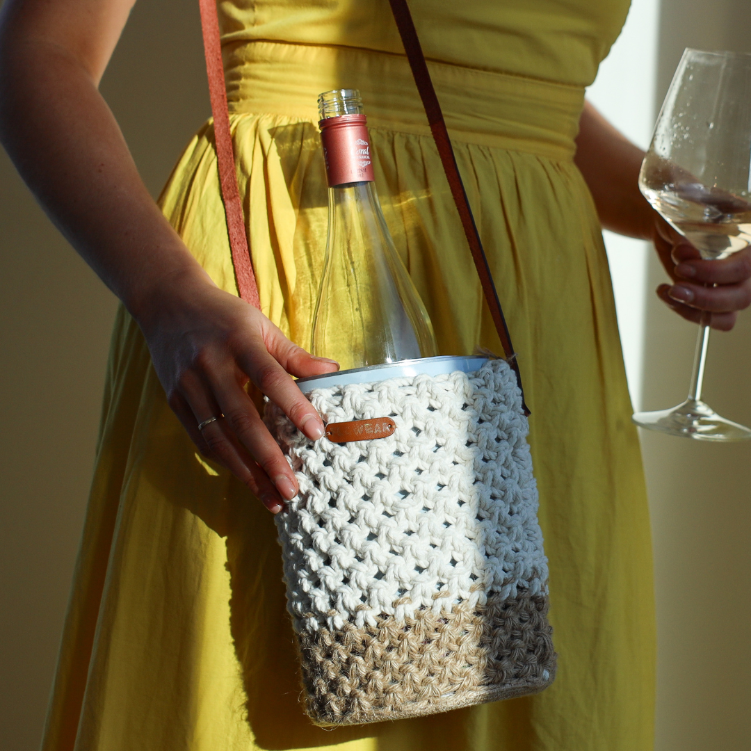 A woman in a yellow dress holding a glass of wine and wearing a withe and beige crochet bag with leather straps with a coolenator wine cooler and bottle of wine in it.