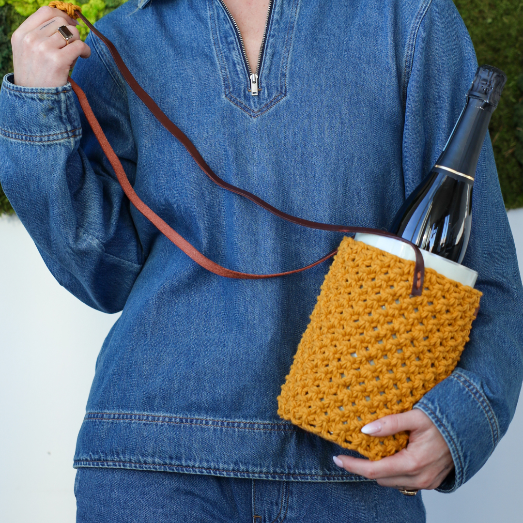 A person wearing all demin holding a coolenator wine cooler in a special crochet yellow back with leather straps.